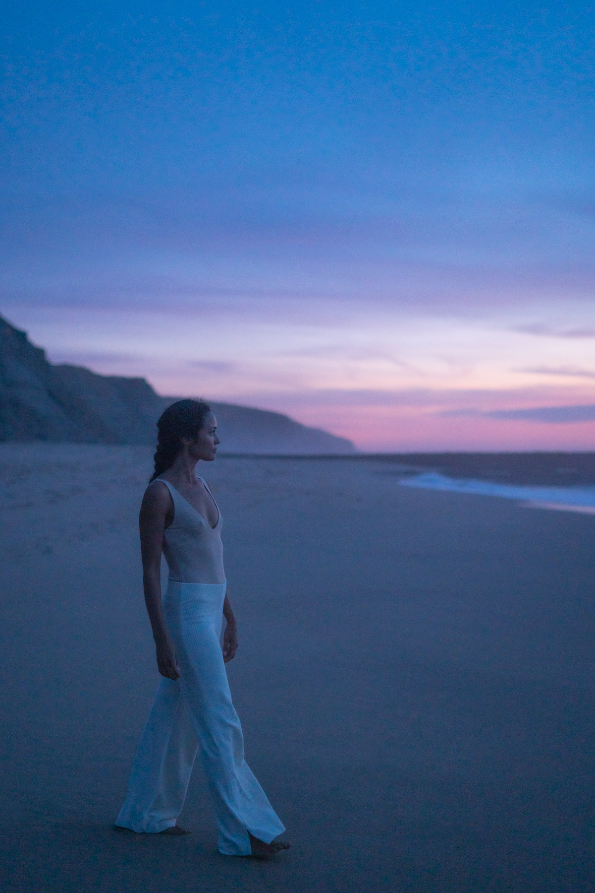 photographie de mode, par Manifa N'Diaye pour le studio magique et Maison la lune de chloé Bloom, représentant une mannequin femme brune en body rose sur une plage de ericiera Portugal, avec un coucher de soleil et des vagues en arrière plan